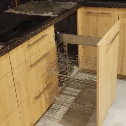 Wysuwana metalowa półka cargo w szafce kuchennej z jasnego drewna, widoczny mechanizm wysuwania, blat z ciemnego, nakrapianego kamienia.