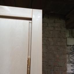 SŁAWOMIR WĘGRZYN SŁAWEX USŁUGI OGÓLNOSTOLATRSKIE - Fragment skrzydła drzwiowego z zawiasem w kolorze złotym, widoczne na tle surowej, ceglanej ściany i drewnianego stropu w trakcie montażu.