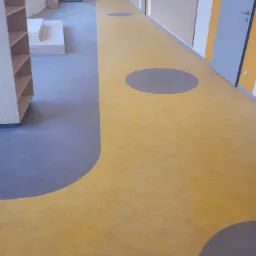 Nowoczesna podłoga w placówce edukacyjnej z żółtą wykładziną i szarymi okręgami, widok na korytarz z jasnymi ścianami i drewnianymi elementami, perspektywa od dołu.