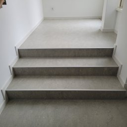 PATRICK KOWALSKI - Schody wykończone szarą wykładziną z delikatnym wzorem, widok z dołu na trzy stopnie z aluminiowymi listwami krawędziowymi, białe ściany i listwy przypodłogowe.