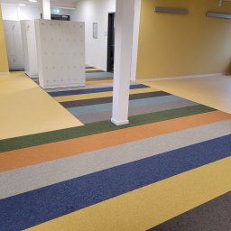 PATRICK KOWALSKI - Wnętrze korytarza z ułożoną wielobarwną wykładziną dywanową w pasy: żółty, niebieski, szary, pomarańczowy i zielony, z białymi szafkami i kolumną w tle.