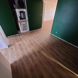 PATRICK KOWALSKI - Pokój z nową, jasnobrązową podłogą imitującą drewno; na ścianach ciemnozielona farba; w otwartym schowku ustawione w kartonach pralki.