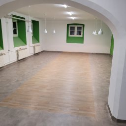 PATRICK KOWALSKI - Jasne, przestronne wnętrze z częściowo położoną wykładziną imitującą drewno, widok przez łukowate przejście z białymi ścianami i zielonymi wnękami okiennymi.