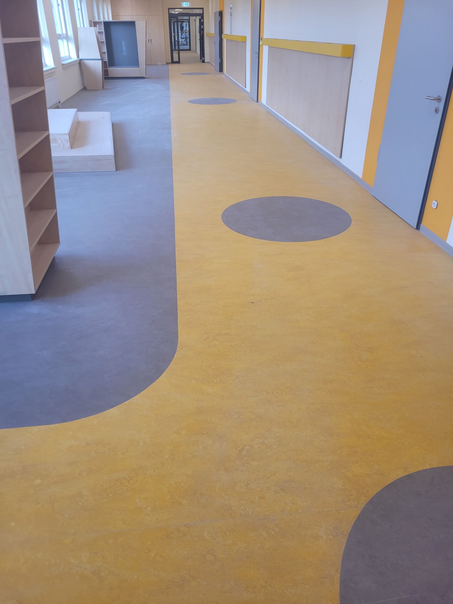 Nowoczesna podłoga w placówce edukacyjnej z żółtą wykładziną i szarymi okręgami, widok na korytarz z jasnymi ścianami i drewnianymi elementami, perspektywa od dołu.