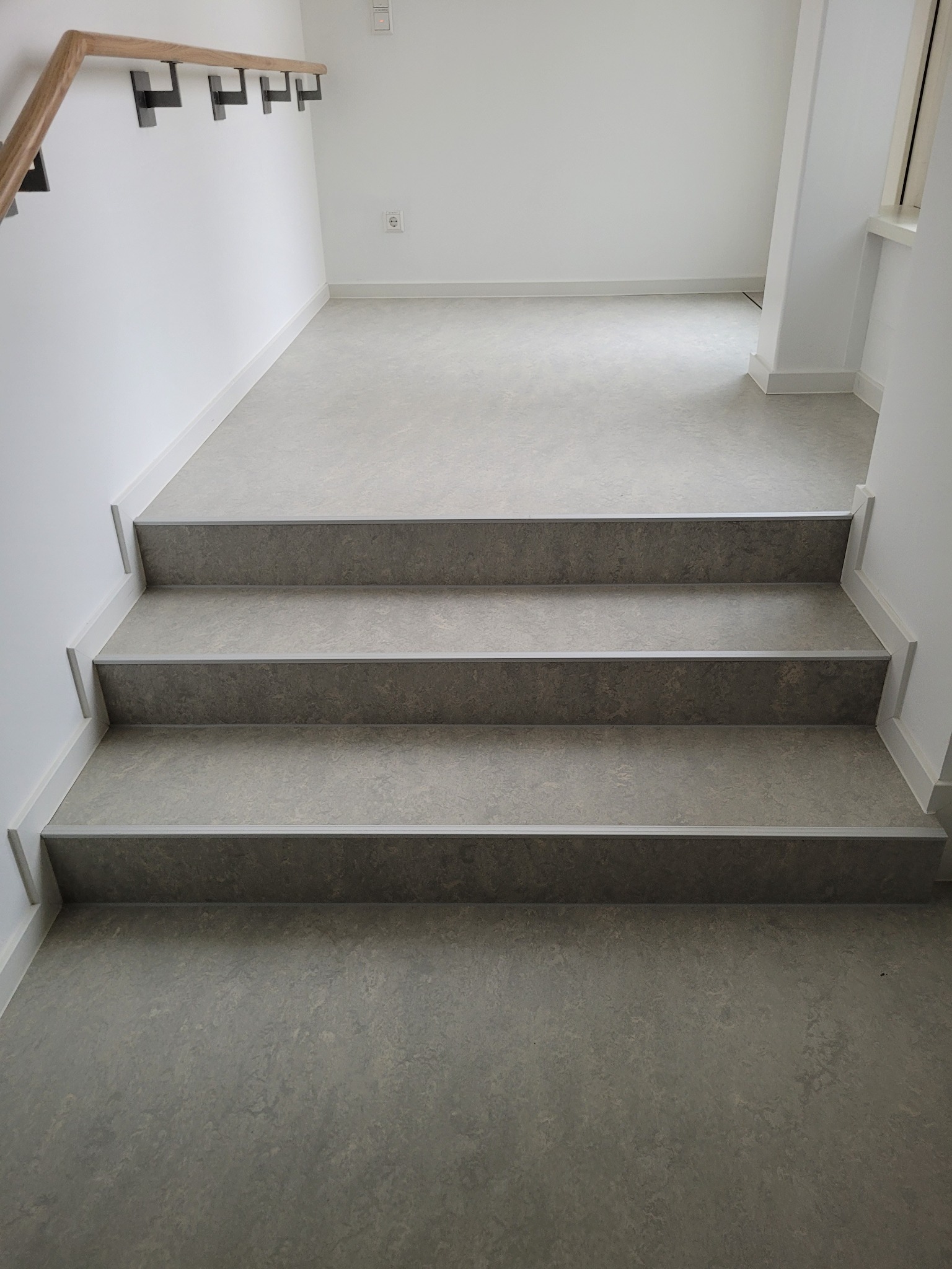 Schody wykończone szarą wykładziną z delikatnym wzorem, widok z dołu na trzy stopnie z aluminiowymi listwami krawędziowymi, białe ściany i listwy przypodłogowe.