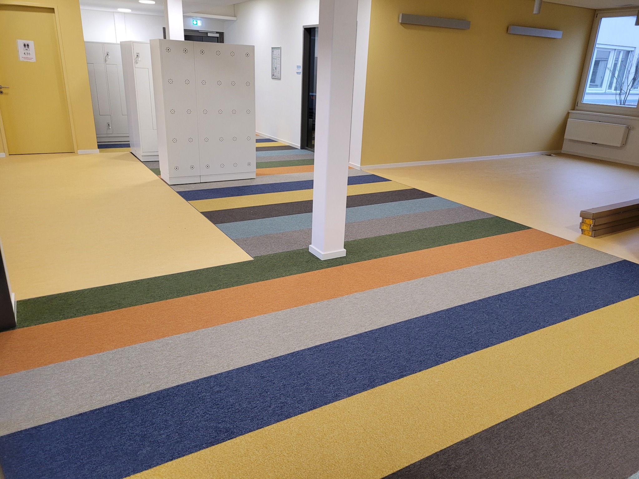 Wnętrze korytarza z ułożoną wielobarwną wykładziną dywanową w pasy: żółty, niebieski, szary, pomarańczowy i zielony, z białymi szafkami i kolumną w tle.