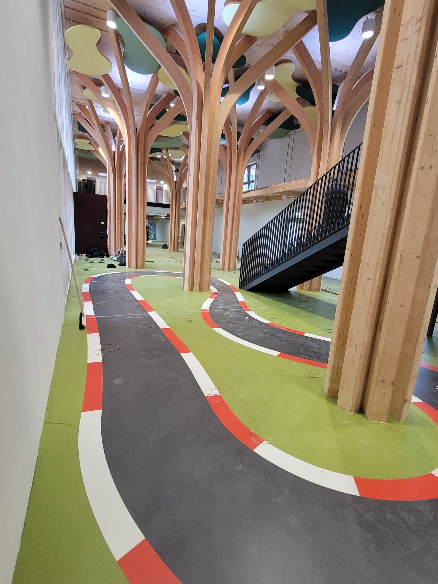 Wnętrze sali zabaw z drewnianymi kolumnami imitującymi drzewa i podłogą wyłożoną wykładziną w kolorze zielonym z namalowanym torem wyścigowym w kolorach szarym, białym i czerwonym.