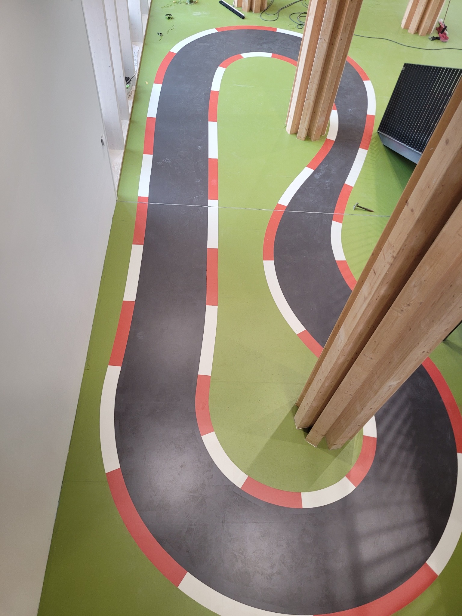 Widok z góry na zieloną podłogę z namalowanym szarym torem wyścigowym z czerwono-białymi krawężnikami, przeciętym przez drewniane słupy konstrukcyjne