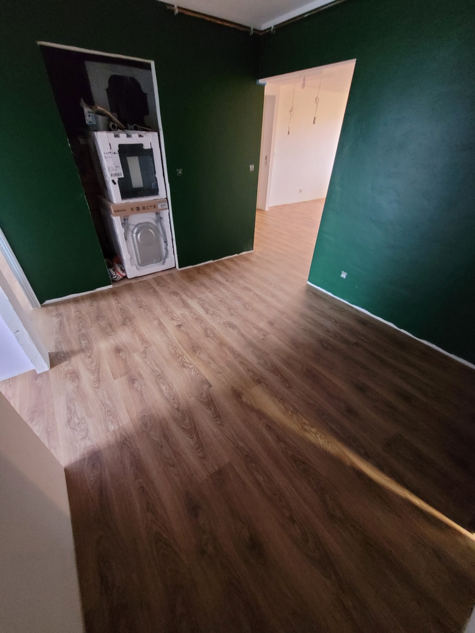 Pokój z nową, jasnobrązową podłogą imitującą drewno; na ścianach ciemnozielona farba; w otwartym schowku ustawione w kartonach pralki.