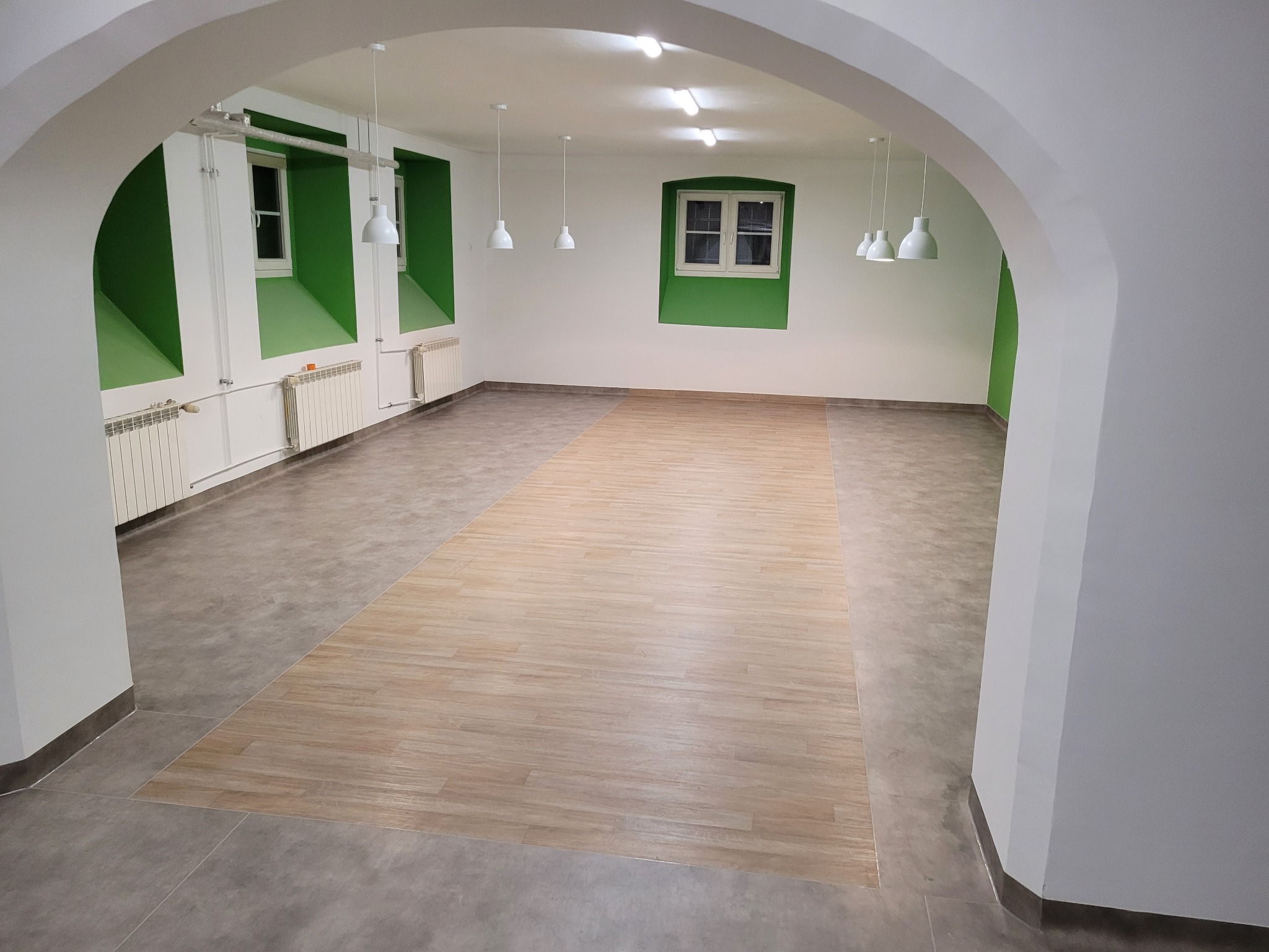 Jasne, przestronne wnętrze z częściowo położoną wykładziną imitującą drewno, widok przez łukowate przejście z białymi ścianami i zielonymi wnękami okiennymi.