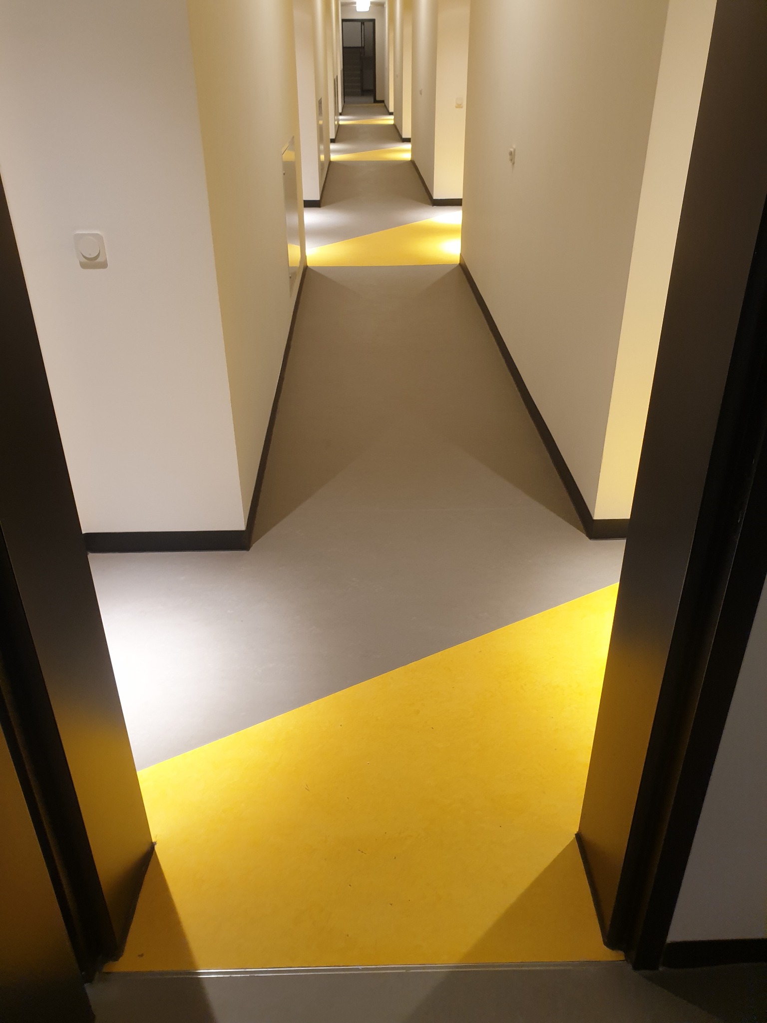 Długi korytarz z geometrycznym wzorem z szarego i żółtego linoleum, widok zza czarnej futryny drzwi.