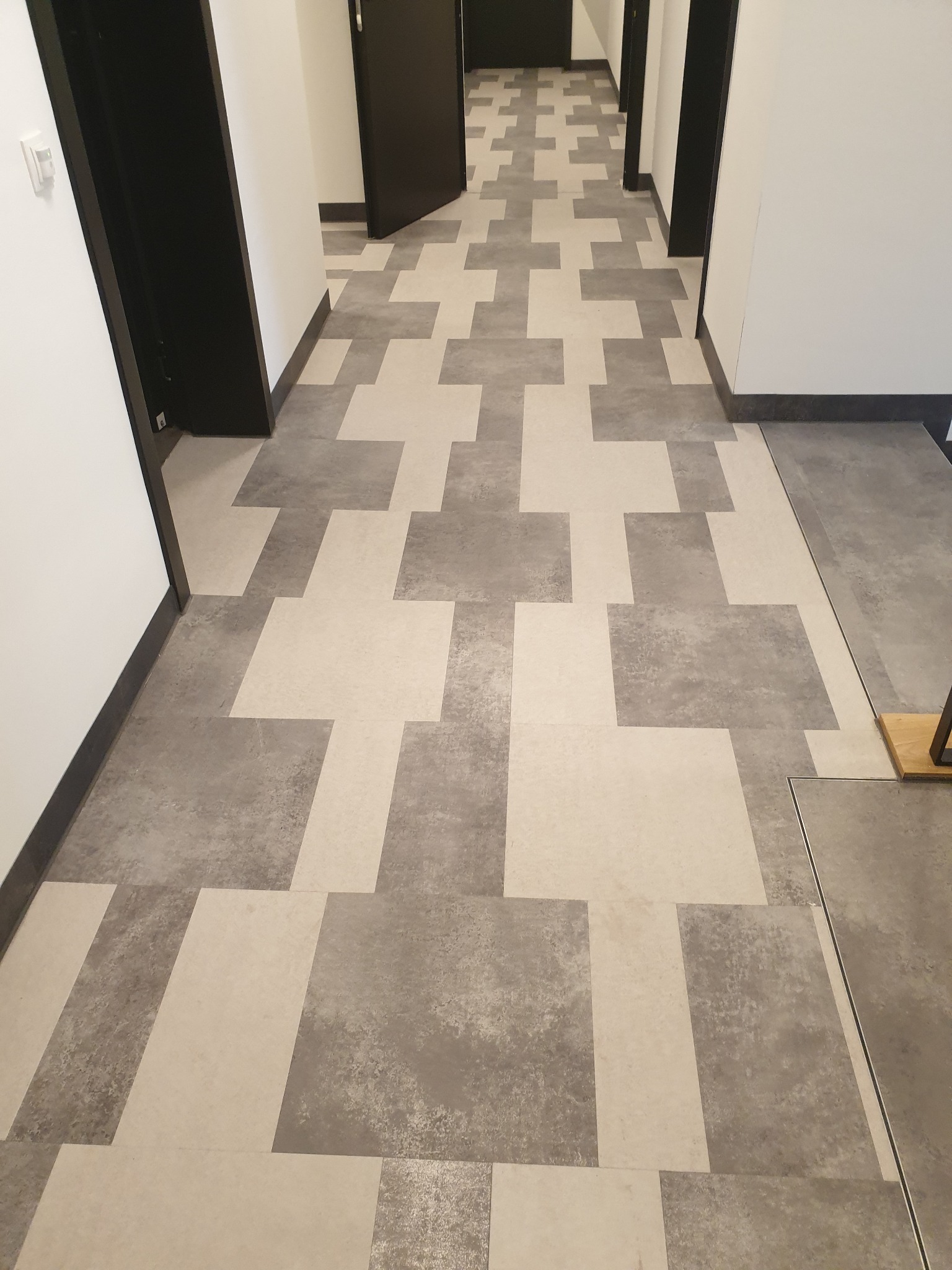 Wzór podłogi w korytarzu ułożony z prostokątnych płytek w odcieniach szarości i beżu, tworzących abstrakcyjny motyw krzyża.
