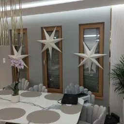 Jadalnia z marmurowym stołem, szarymi krzesłami w kształcie muszli, drewnianymi oknami z żaluzjami oraz dekoracyjnymi gwiazdami na ścianie i złotymi lampami wiszącymi.