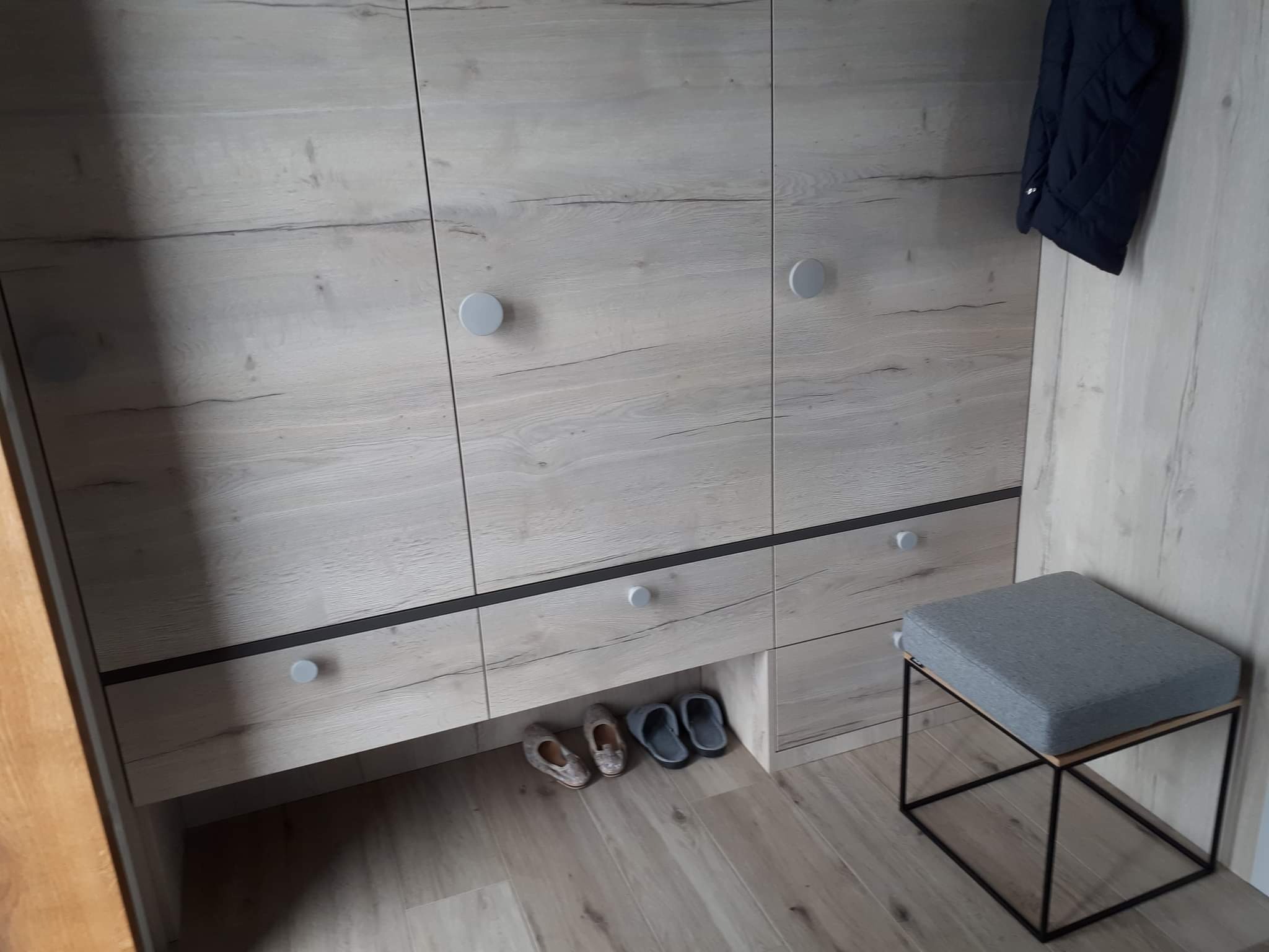 Jasna szafa z drewnopodobnymi frontami, okrągłe uchwyty, szuflady z czarnymi akcentami, damskie pantofle pod szafką, szary tapicerowany stołek oraz wisząca kurtka.