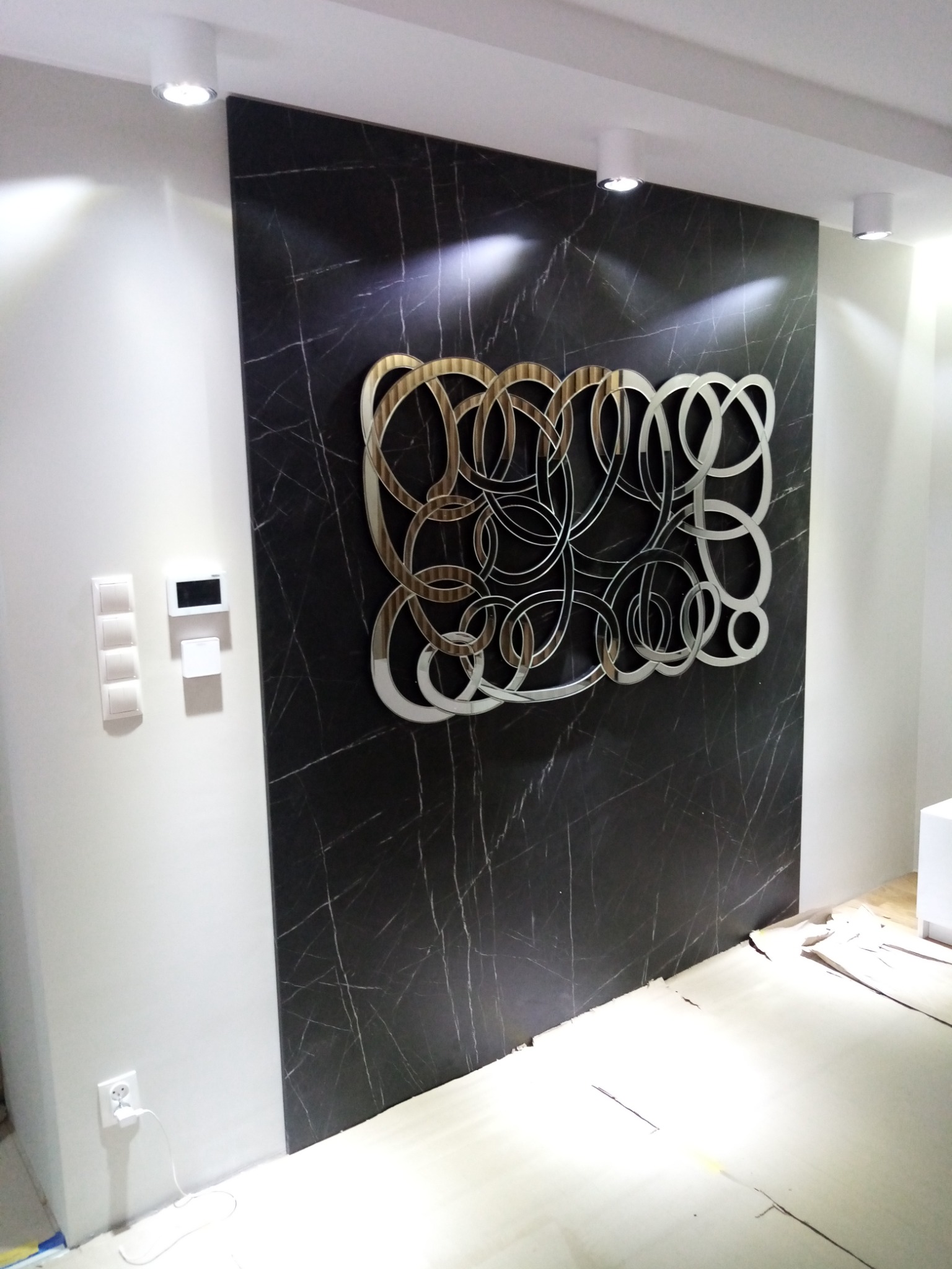 Ściana wykończona czarnym marmurem z dekoracyjną metalową aplikacją w kształcie splecionych okręgów, oświetlona punktowymi lampami sufitowymi, widoczne gniazdko elektryczne i panel sterowania...