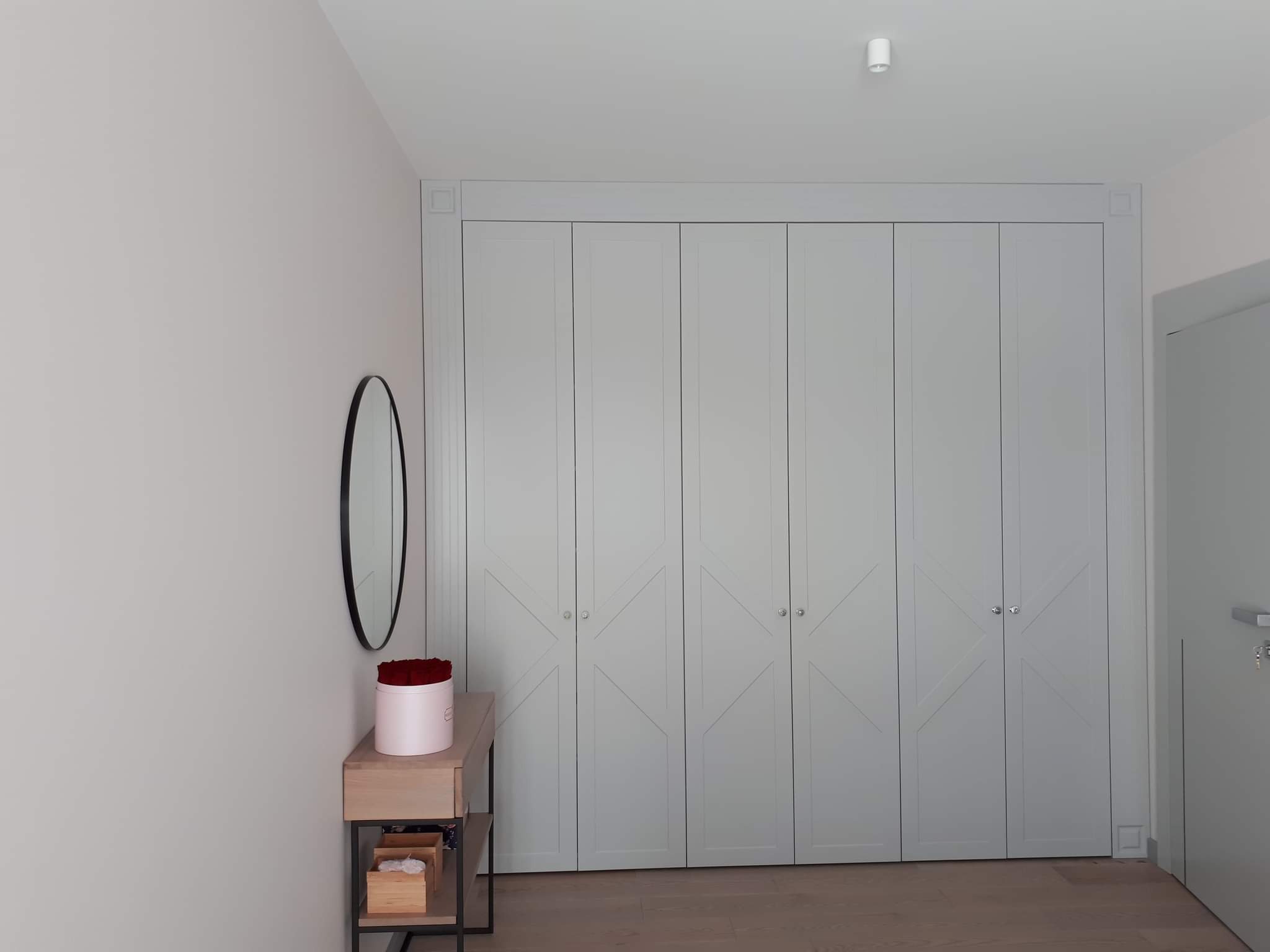 Jasnoszara szafa wnękowa z sześcioma drzwiami, dekoracyjne krzyże na frontach, owalne lustro i stolik z różami w pudełku, jasna podłoga.