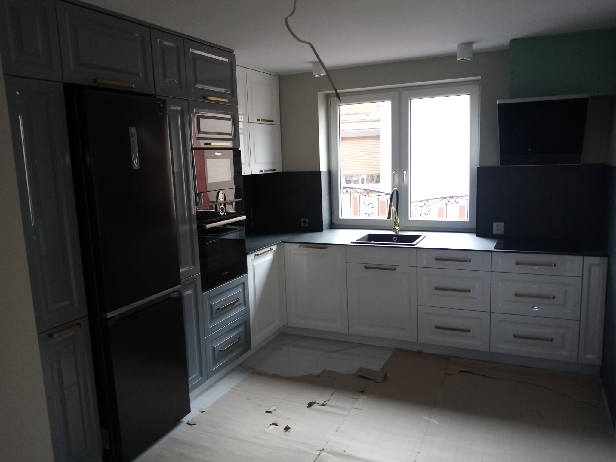 Montaż kuchni: szaro-białe szafki z złotymi uchwytami, czarny blat i lodówka w zabudowie, widoczne zabezpieczenia podłogi kartonem.