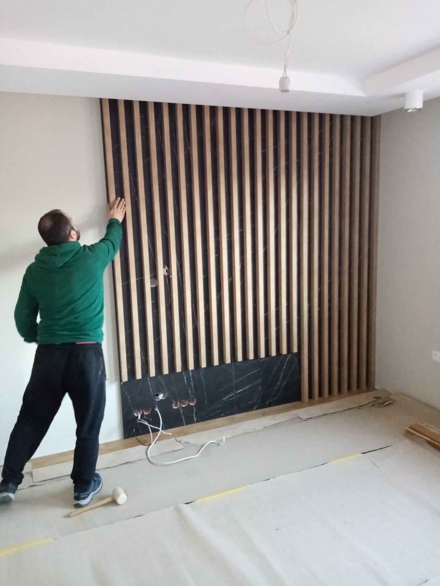 Montaż drewnianych lameli ściennych z elementem dekoracyjnym z czarnego marmuru, widoczny instalator w zielonej bluzie i zabezpieczona folią podłoga.