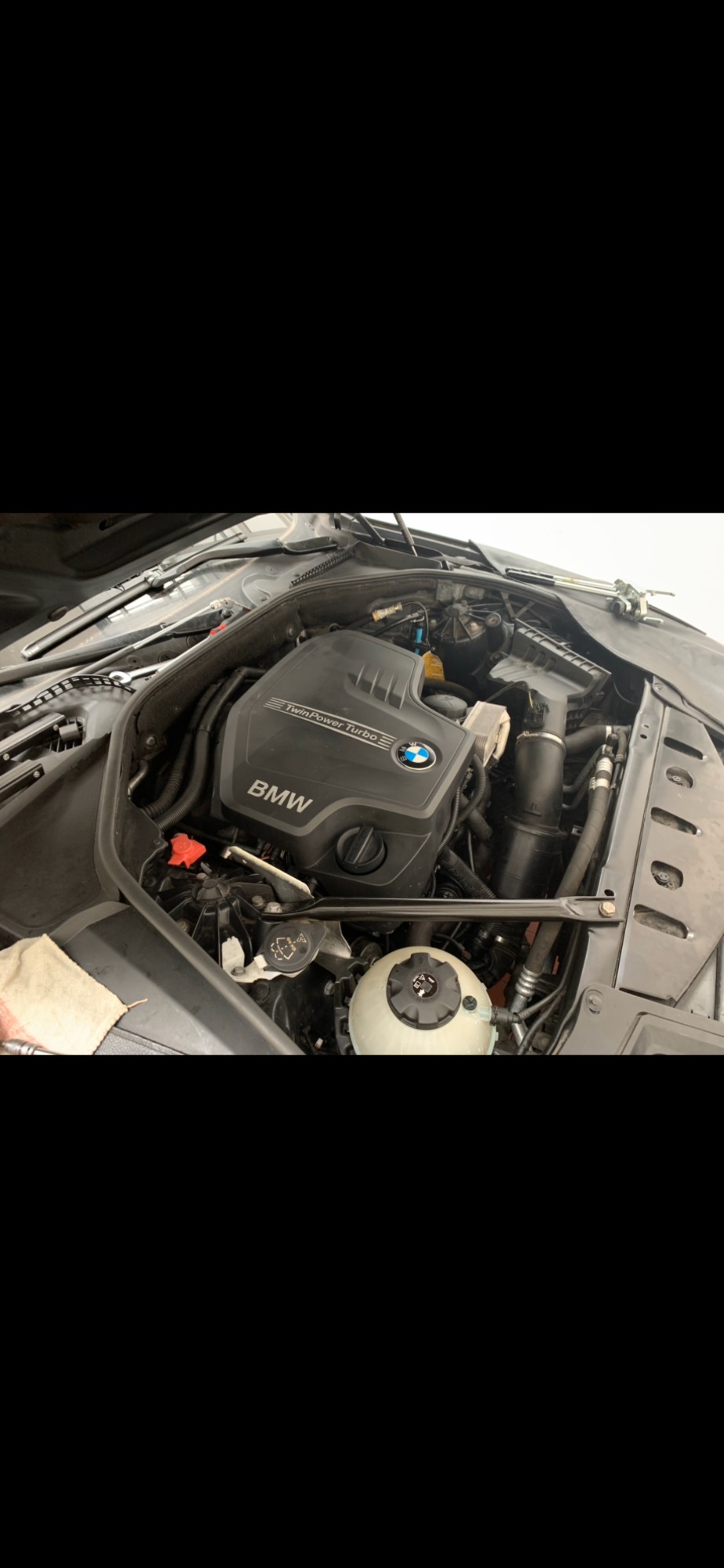 Otwarta maska samochodu BMW, widoczny silnik TwinPower Turbo z elementami osprzętu, w tle biała ściana warsztatu, obok leży szara ściereczka.