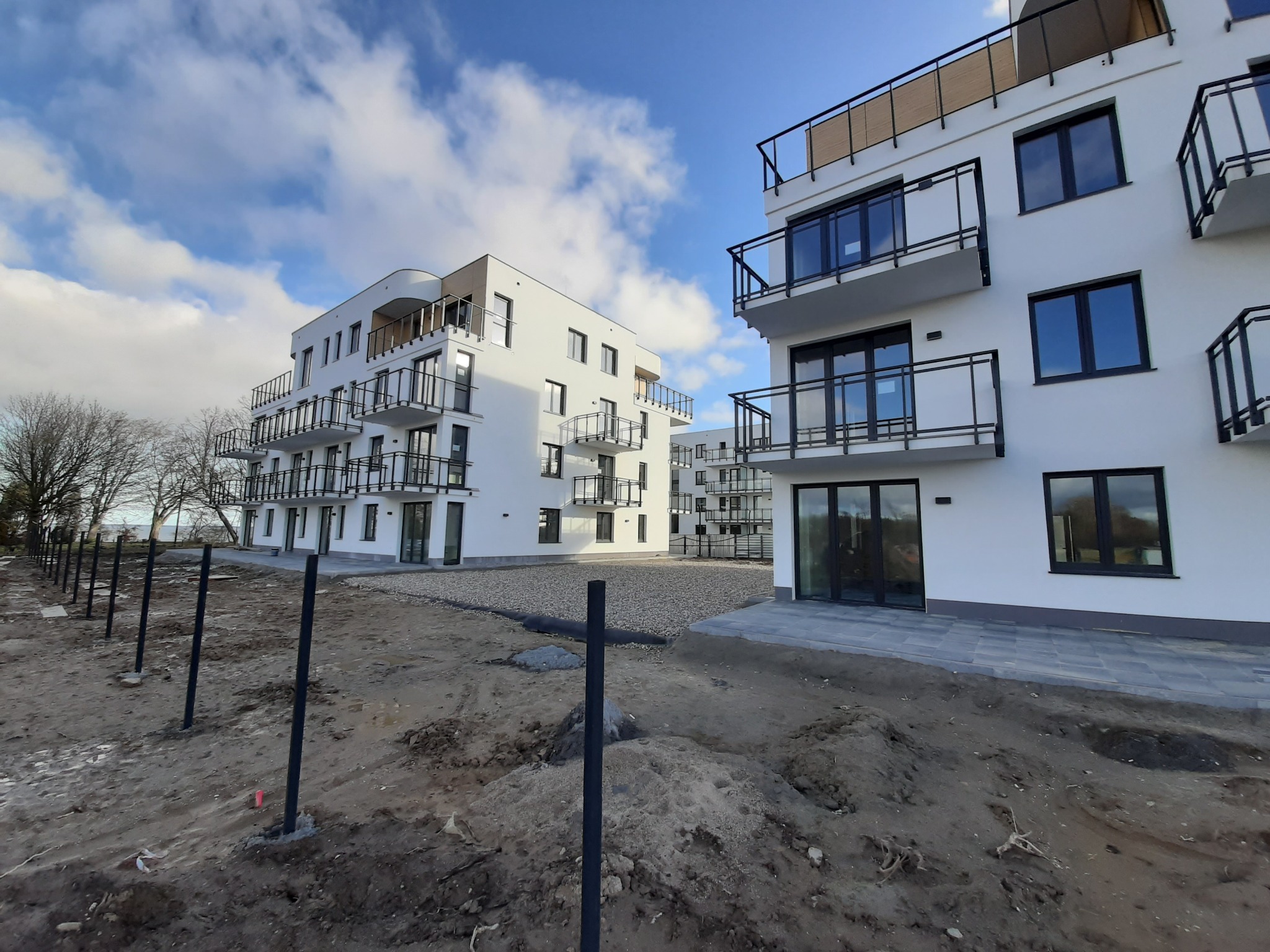 Nowoczesne, białe budynki apartamentowe z balkonami, widok z poziomu gruntu na placu budowy z czarnymi słupkami ogrodzeniowymi, błękitne niebo z chmurami w tle.