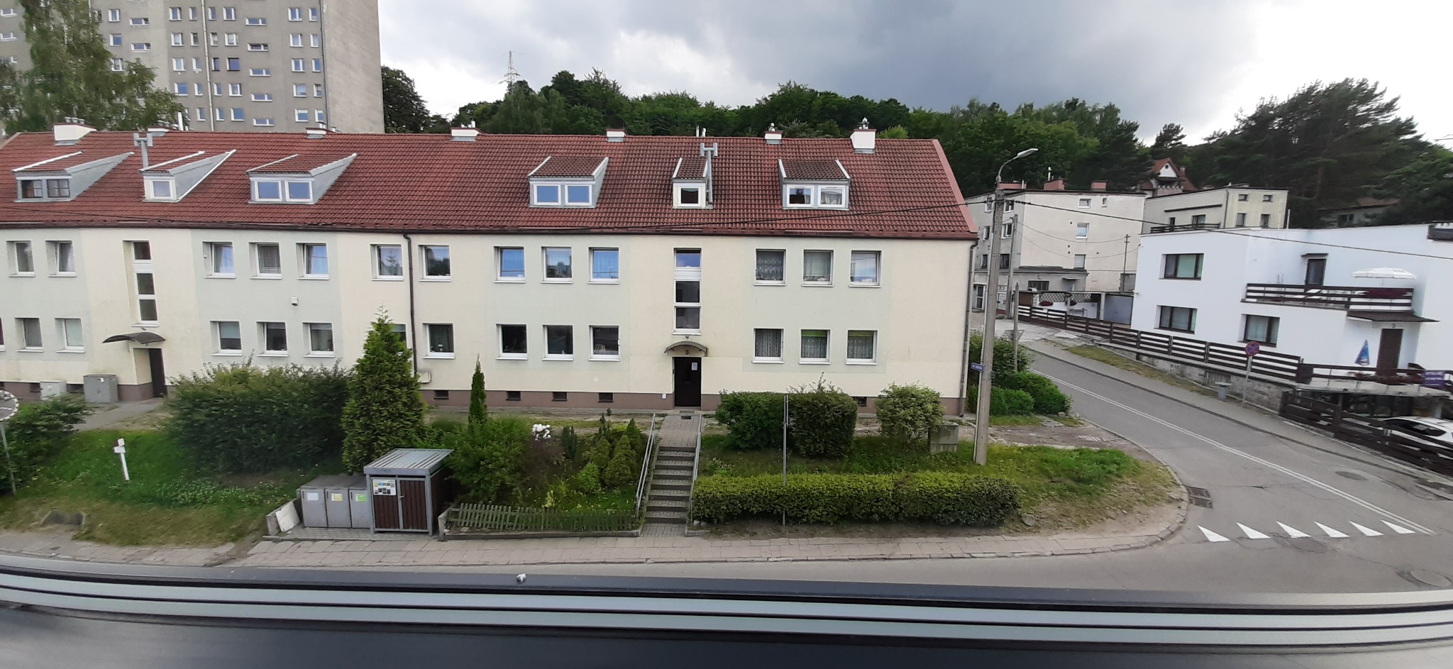 Widok z okna na trzypiętrowy budynek mieszkalny z czerwonym dachem, zadbanym trawnikiem i niewielkim ogrodem przed wejściem, w tle drzewa i pochmurne niebo.