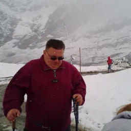 Mężczyzna w bordowej bluzie i okularach przeciwsłonecznych, stojący na ośnieżonej górskiej ścieżce z kijkami trekkingowymi i różańcem w rękach, w tle zamglony krajobraz górski z osobą w czerwonej...