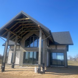 Mariusz Skoczylas BAU-MAX - Surowy stan domu z dachem i oknami, widoczne pustaki i betonowe słupy, w tle błękitne niebo. Dom w trakcie budowy, z elementami konstrukcyjnymi na zewnątrz.