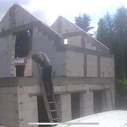 Mariusz Skoczylas BAU-MAX - Budowa domu z betonu komórkowego: murarz na drabinie, ściany w trakcie wznoszenia, widoczne zbrojenia i elementy konstrukcyjne. Tło: zieleń drzew.