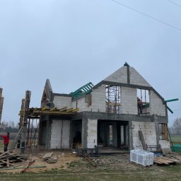 Mariusz Skoczylas BAU-MAX - Budowa domu z białych bloczków, widoczne szalunki, zbrojenia i pracownicy. Surowy stan, zielone elementy dachu, pochmurne niebo w tle. Etap murowania ścian.