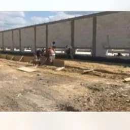 Budowa hali: pracownicy w trakcie murowania ścian z bloczków betonowych. Widoczne szalunki fundamentowe i przygotowany teren pod budowę. Częściowo wzniesiona konstrukcja z otworami okiennymi.