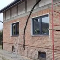 Budynek w trakcie ocieplania fundamentów, widoczne szare płyty izolacyjne na ścianach, czarna rura odprowadzająca wodę i rusztowanie.
