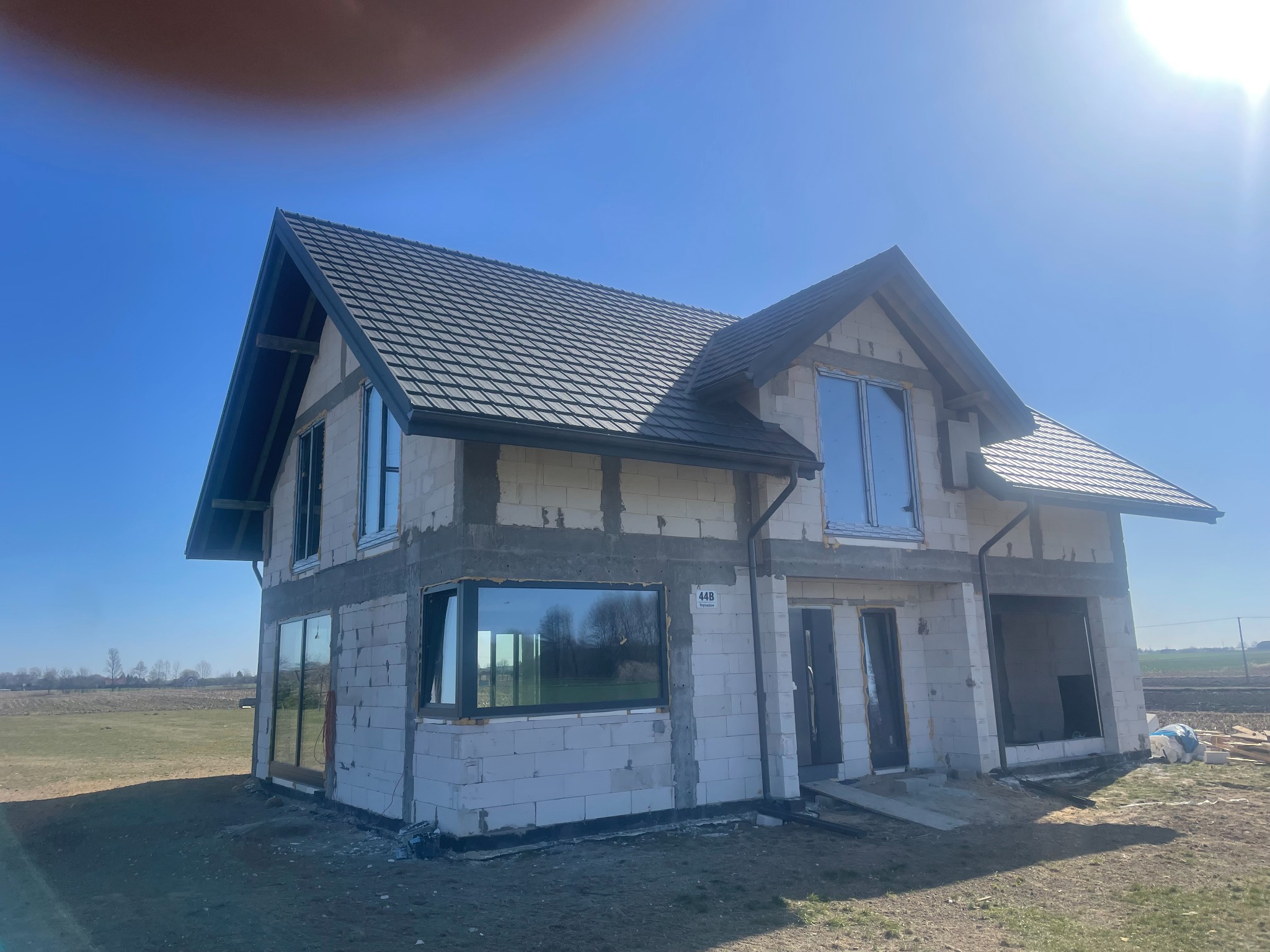 Surowy dom z oknami w Cycowie, w trakcie budowy. Widoczne bloczki, dach z dachówki i zamontowane okna. Jasne niebo w tle, budynek w stanie deweloperskim.