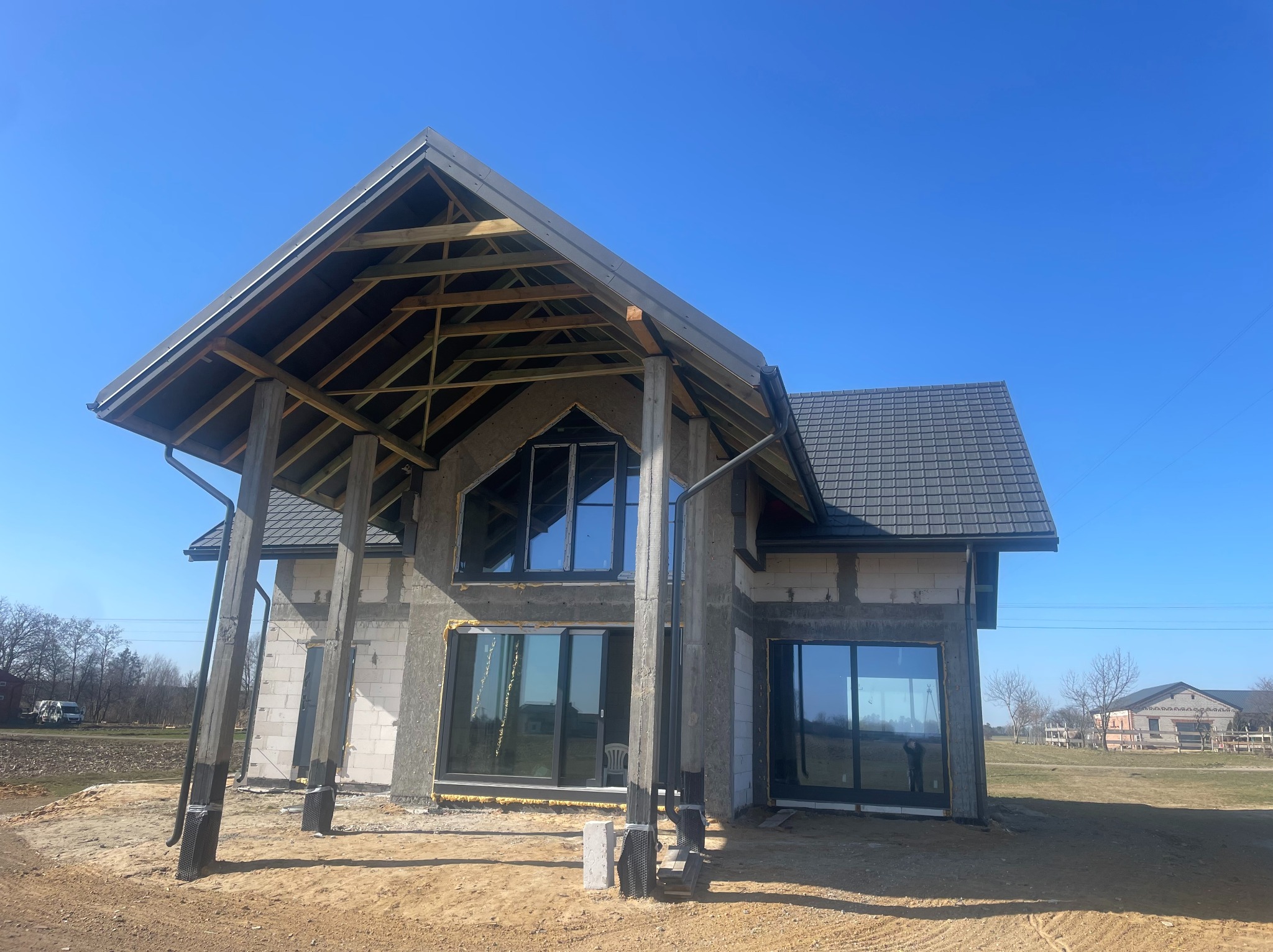 Surowy stan domu z dachem i oknami, widoczne pustaki i betonowe słupy, w tle błękitne niebo. Dom w trakcie budowy, z elementami konstrukcyjnymi na zewnątrz.