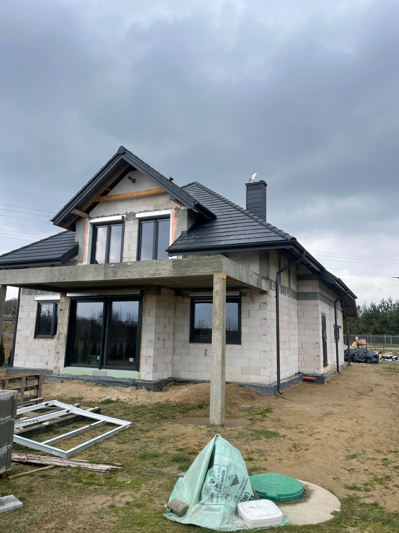 Niedokończony dom z czarną dachówką i oknami, widoczne elementy konstrukcyjne, betonowy balkon, teren budowy z materiałami. Pochmurne niebo w tle.