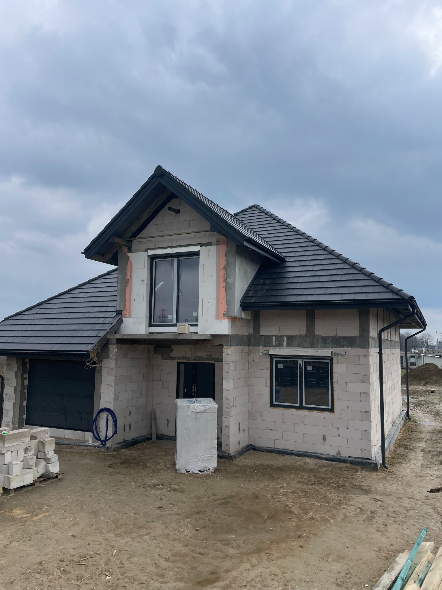 Surowy dom z czarną dachówką w trakcie budowy, widoczne bloczki, okna z czarnymi ramami i materiały budowlane na placu. Pochmurne niebo w tle.