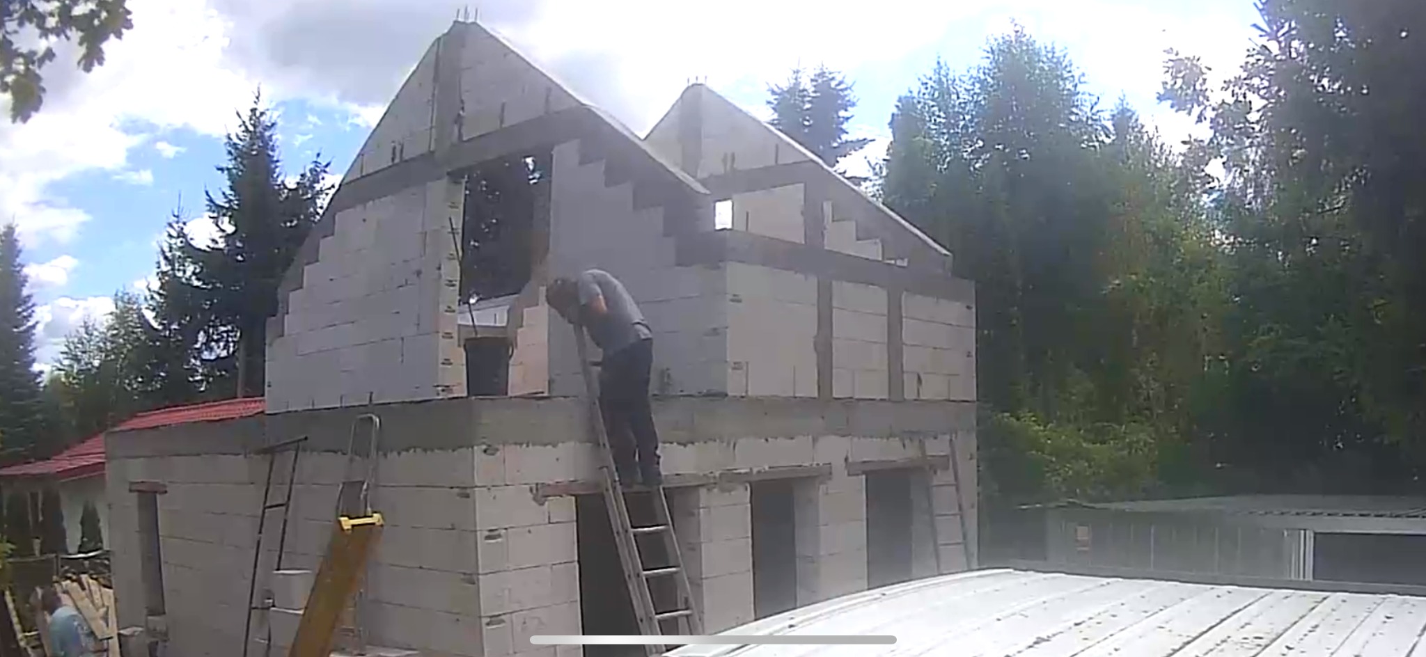 Budowa domu z betonu komórkowego: murarz na drabinie, ściany w trakcie wznoszenia, widoczne zbrojenia i elementy konstrukcyjne. Tło: zieleń drzew.