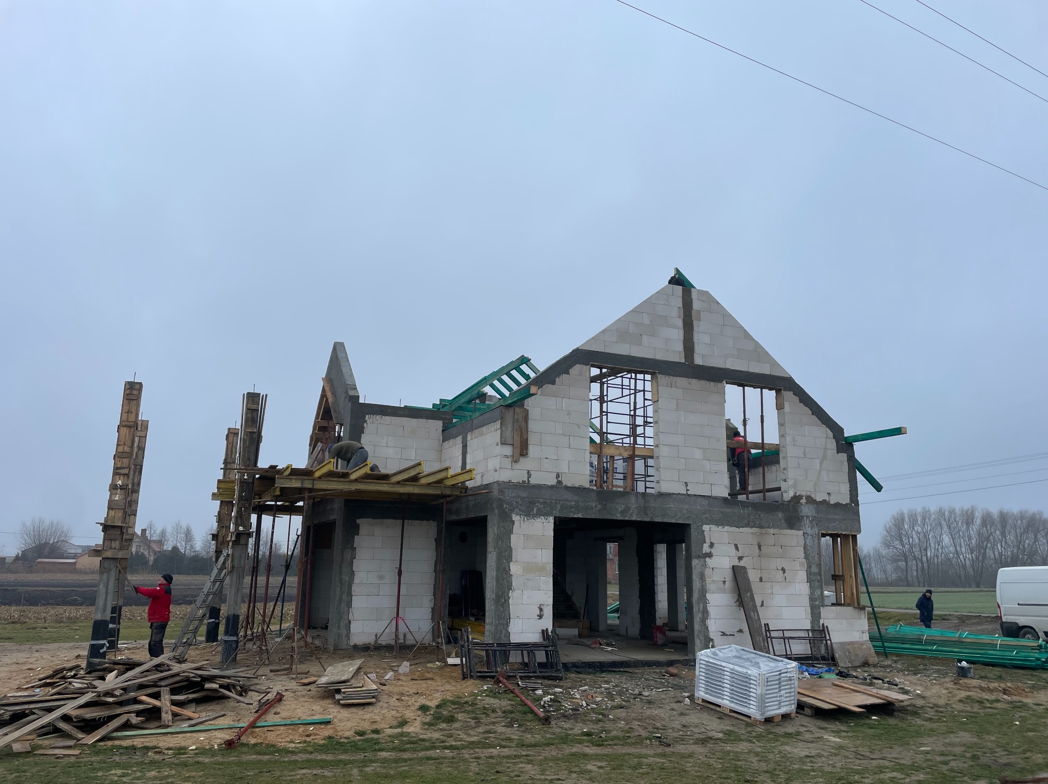 Budowa domu z białych bloczków, widoczne szalunki, zbrojenia i pracownicy. Surowy stan, zielone elementy dachu, pochmurne niebo w tle. Etap murowania ścian.