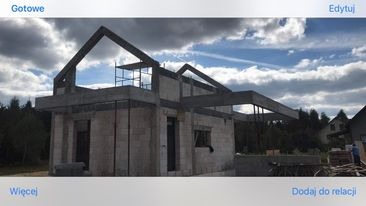 Surowy stan domu murowanego w trakcie budowy, widoczne ściany z betonu komórkowego, stalowe belki stropowe i konstrukcja dachu, rusztowanie, w tle las i pochmurne niebo.