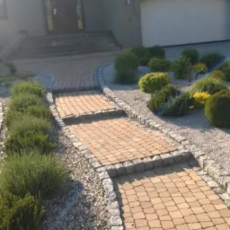 Ogród z ścieżką z kostki brukowej w odcieniach beżu, obrzeża z szarego granitu. Dekoracyjne krzewy i kamień w tle. Podjazd z szarego kamienia.