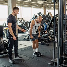 Mariusz Bratek - Trener personalny nadzoruje ćwiczenie na maszynie wyciągowej w siłowni. Instruktor z zegarkiem na ręku obserwuje postępy mężczyzny w białym tank topie.