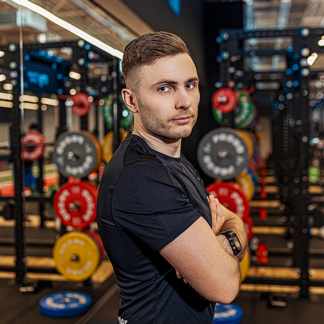Portret trenera personalnego w czarnej koszulce, z zegarkiem na ręku, na tle stojaków z obciążeniami w siłowni. Skrzyżowane ramiona, pewny wzrok.
