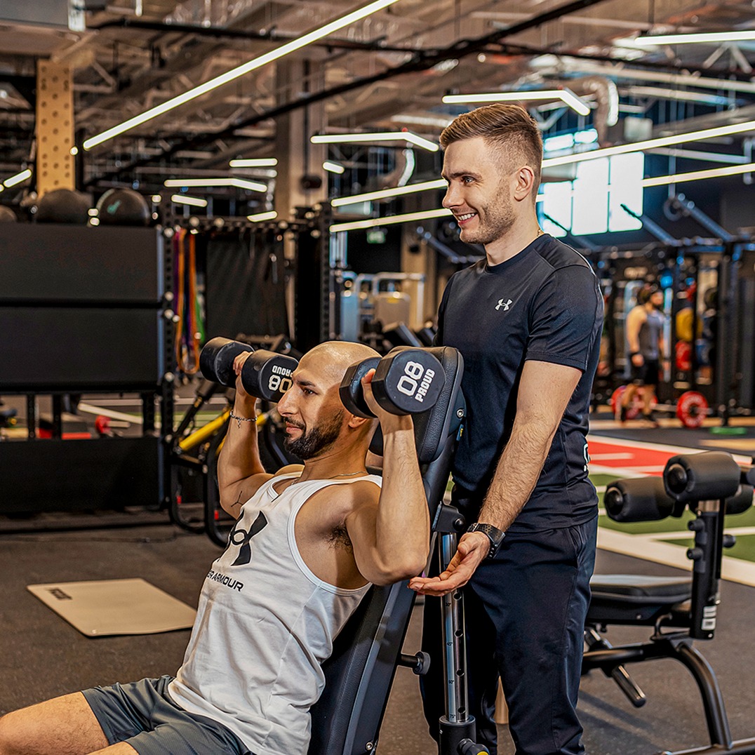 Mężczyzna ćwiczy wyciskanie sztangielek pod okiem uśmiechniętego instruktora w nowoczesnej siłowni z profesjonalnym sprzętem do ćwiczeń.