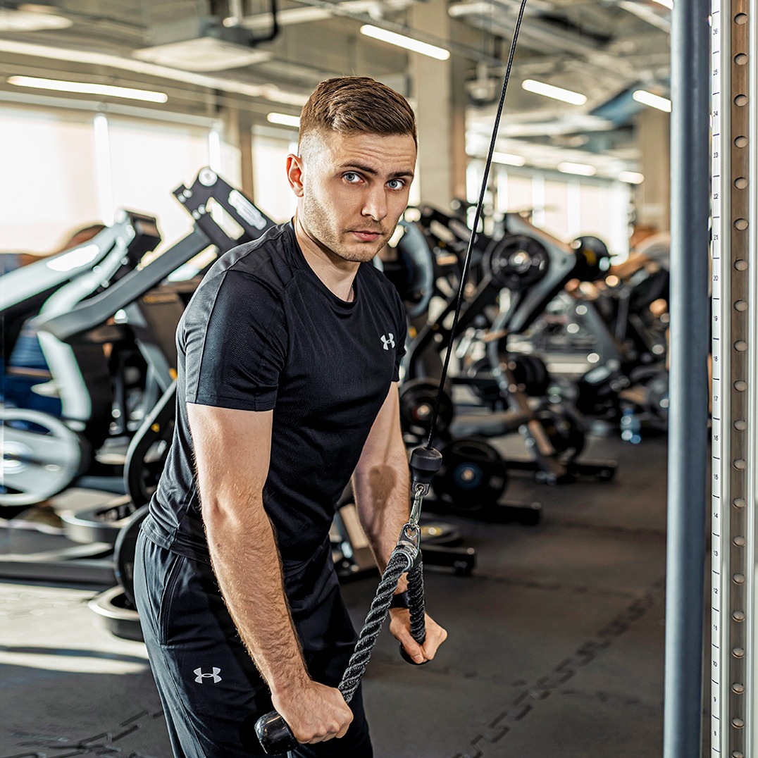 Mężczyzna w czarnym stroju sportowym ćwiczy na maszynie w siłowni, trzymając linę. W tle widoczne inne urządzenia do ćwiczeń i oświetlenie sufitowe.