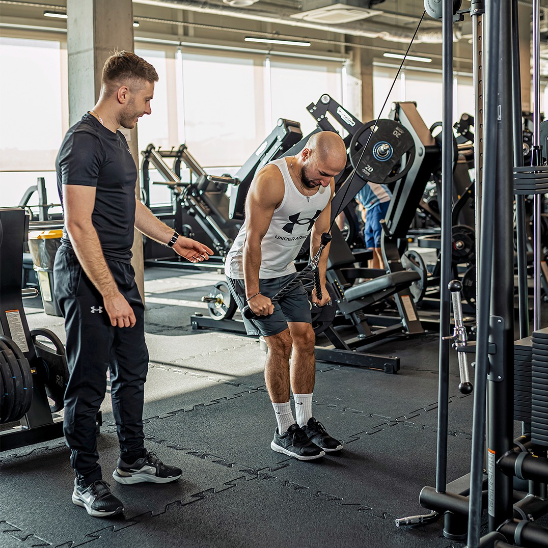 Trener personalny nadzoruje ćwiczenie na maszynie wyciągowej w siłowni. Instruktor z zegarkiem na ręku obserwuje postępy mężczyzny w białym tank topie.