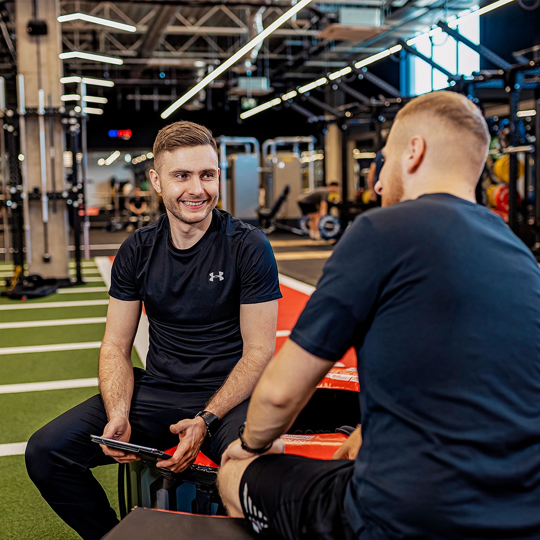 Rozmowa w siłowni: uśmiechnięty mężczyzna z tabletem siedzi naprzeciwko klienta, omawiając plan treningowy na tle profesjonalnego sprzętu.