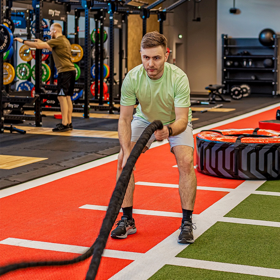 Mężczyzna w sportowym stroju ćwiczy z grubą liną w nowoczesnej siłowni. W tle inny mężczyzna wykonuje ćwiczenia z obciążeniem. Widoczny sprzęt do crossfitu.