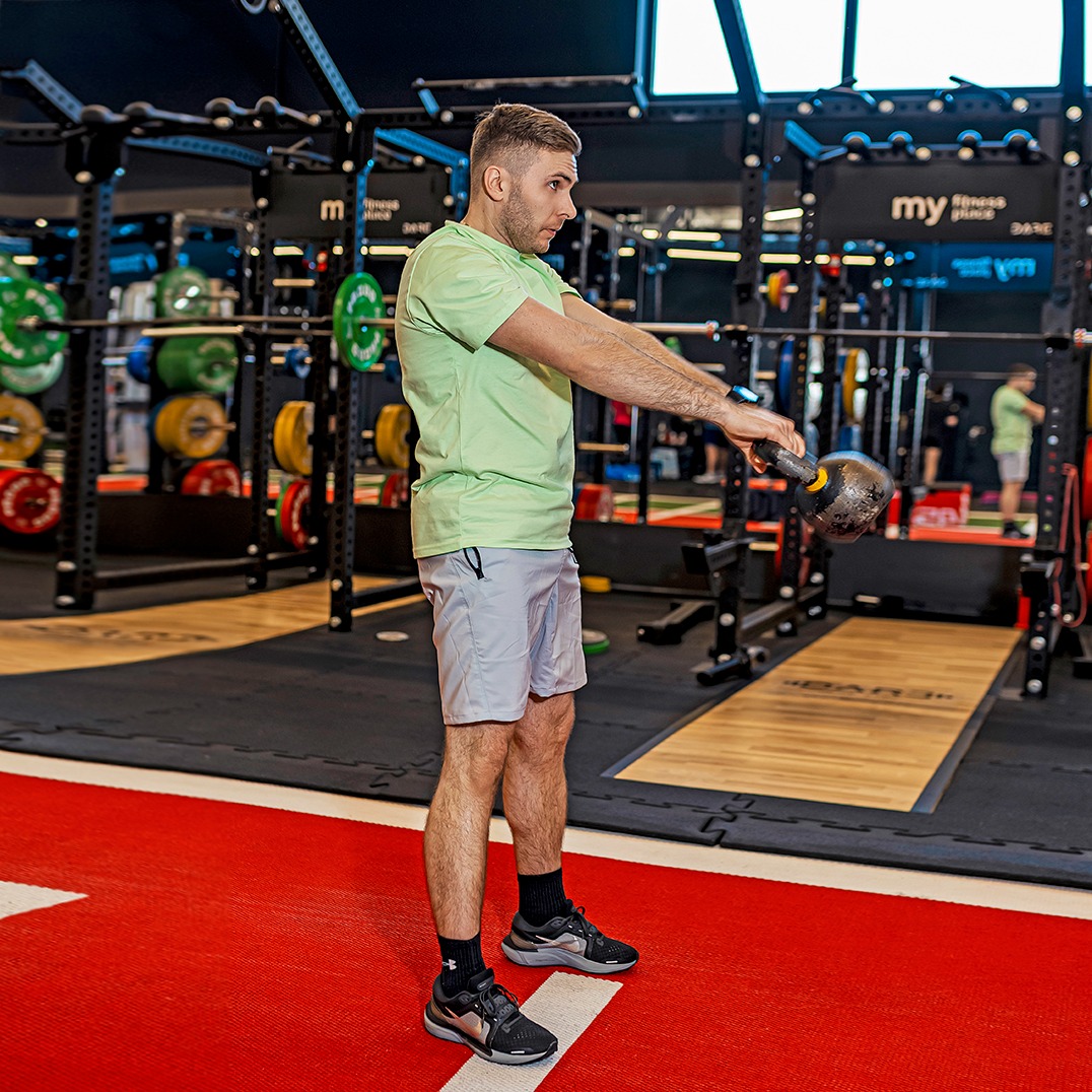 Mężczyzna w sportowym stroju ćwiczy z kettlebellem w siłowni. Widoczne wyposażenie siłowni, czerwona podłoga i lustro z odbiciem ćwiczącego.