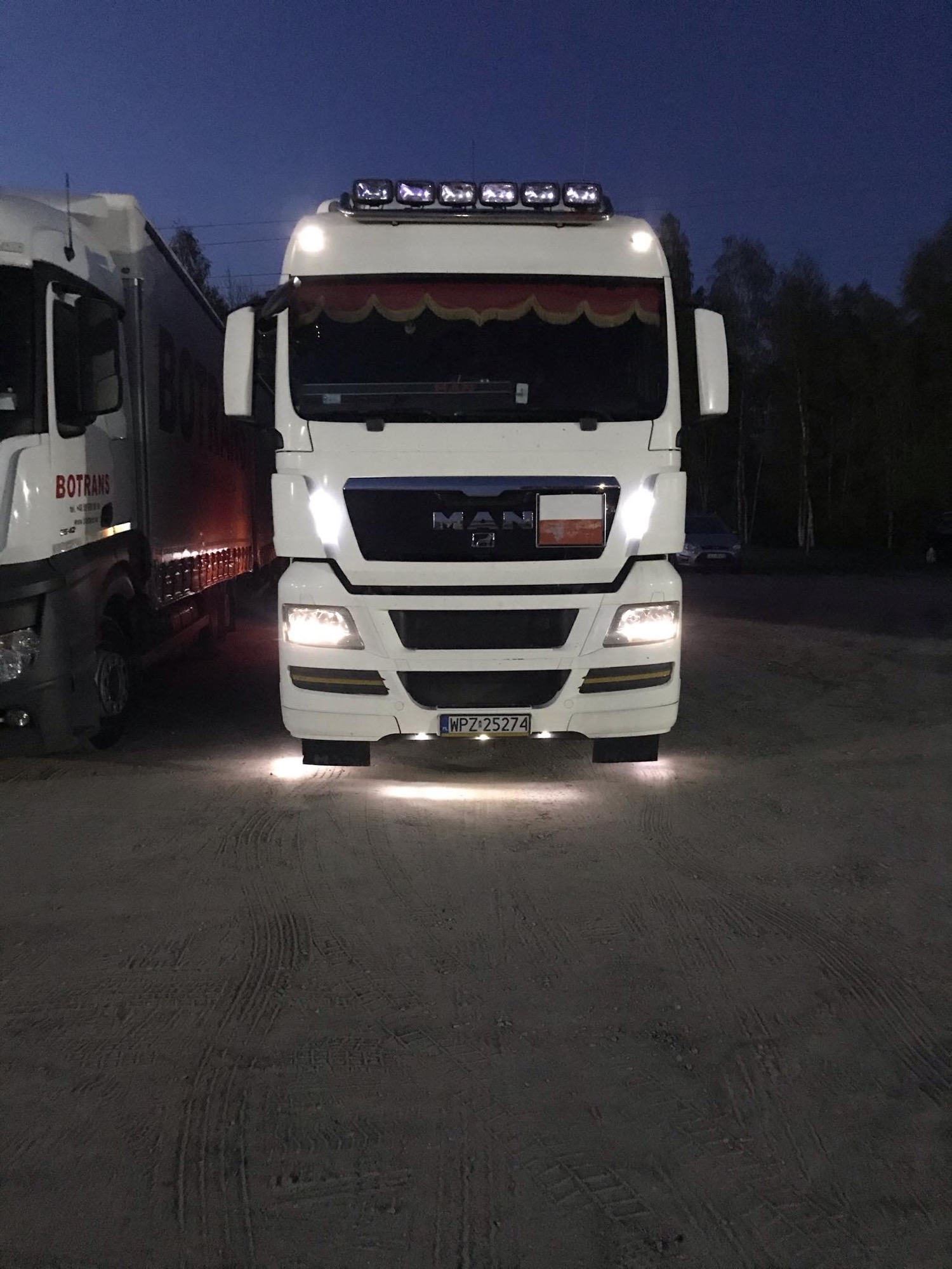 Biała ciężarówka MAN z polską flagą na grillu, oświetlona reflektorami, zaparkowana na żwirowym placu o zmierzchu.