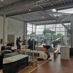 Skinder Global Service Sp.j - Pracownicy w trakcie demontażu i pakowania wyposażenia salonu samochodowego, meble owinięte folią bąbelkową i czarną folią stretch, widoczne witryny szklane, manekiny i szafki, tło stanowi...