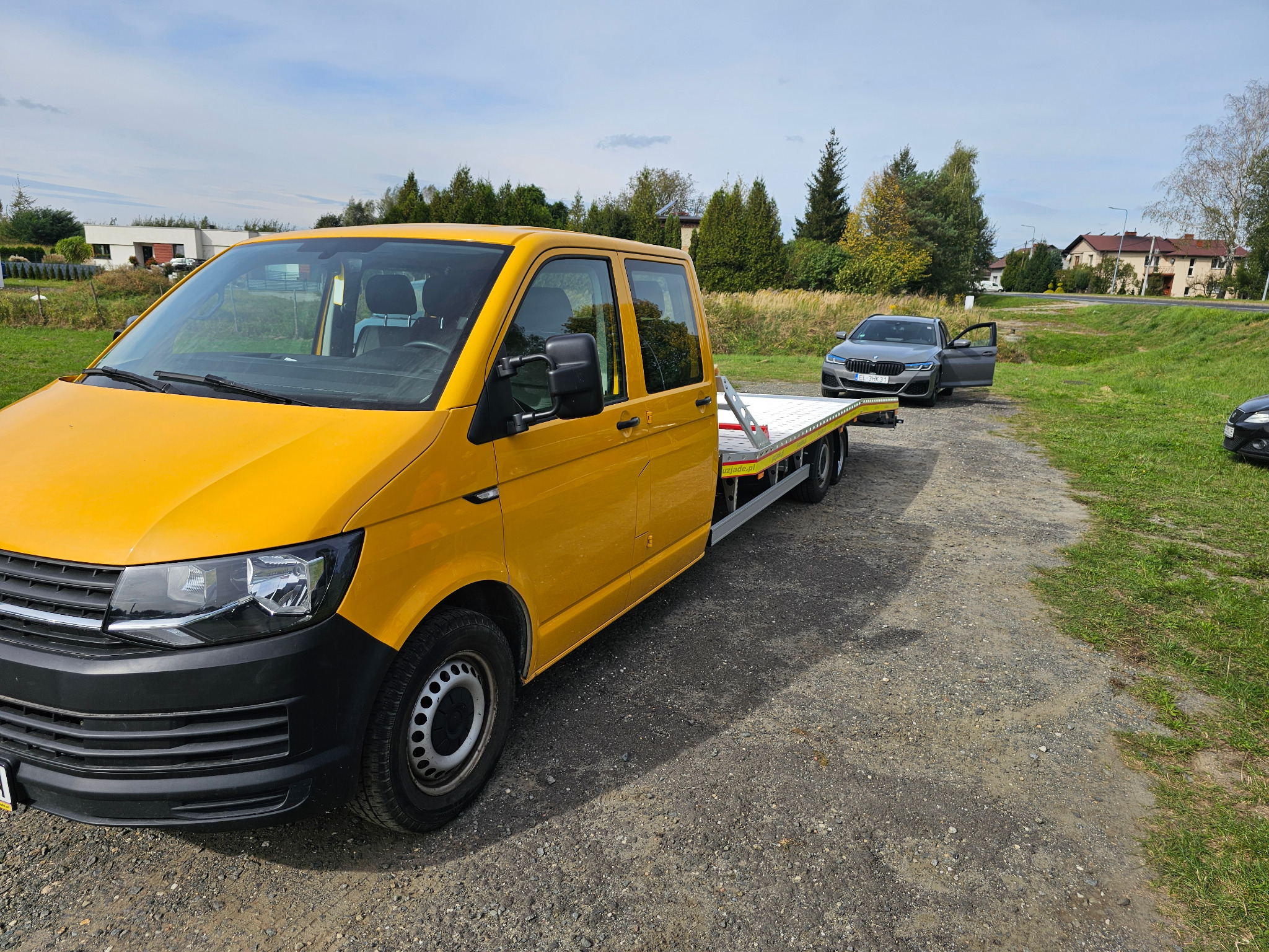 Żółty samochód laweta z platformą załadunkową na żwirowej drodze, obok szary samochód osobowy z otwartymi drzwiami kierowcy.